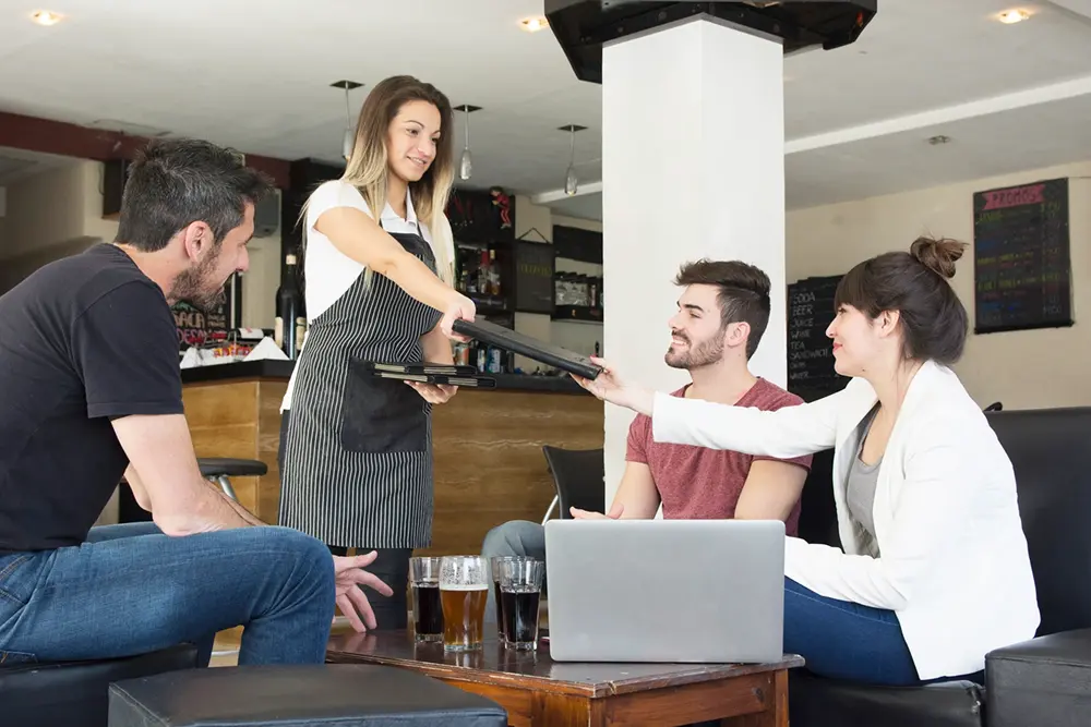Waitress presenting menu to female customer with friends at the bar.