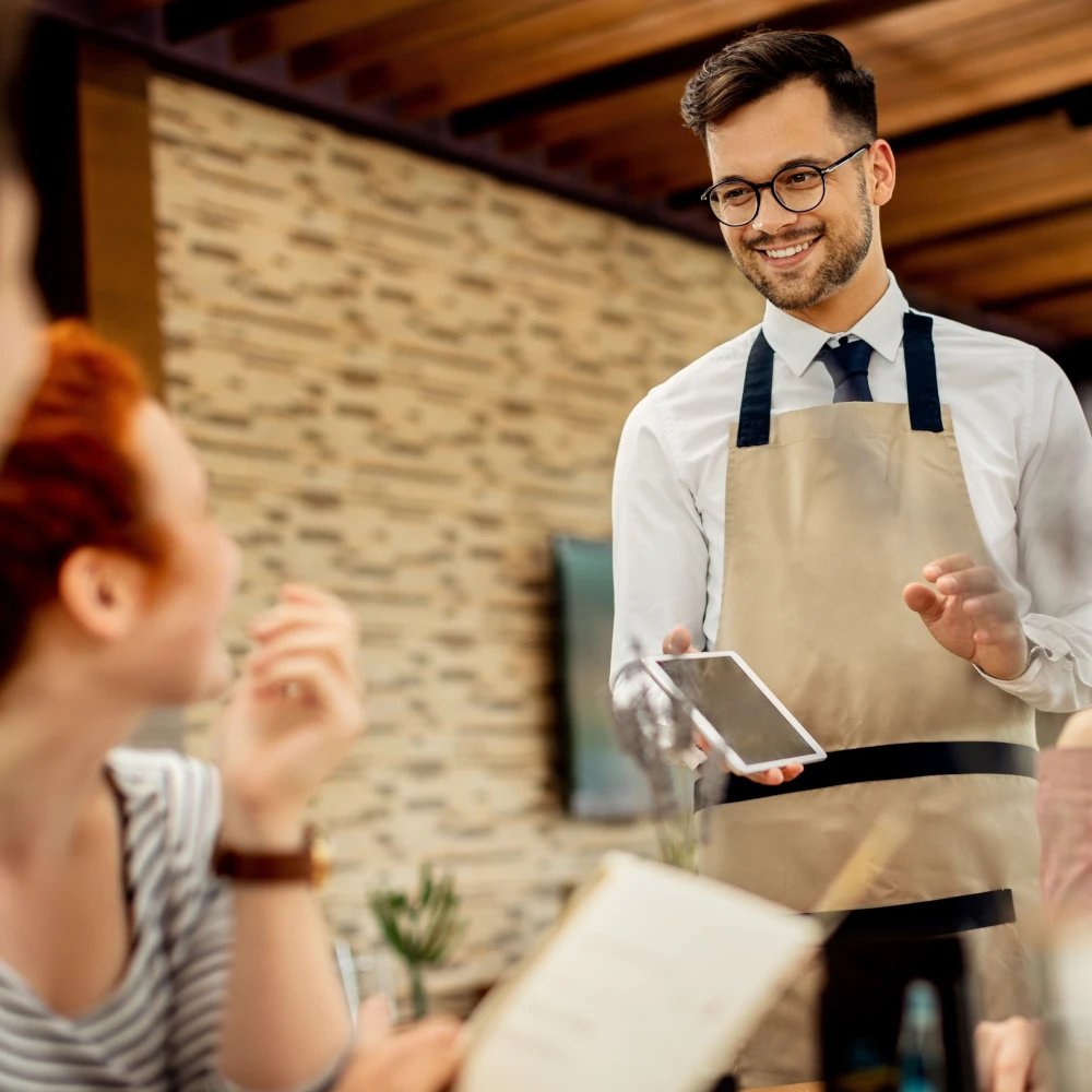 A smiling male waiter chatting to customers at a restaurant table